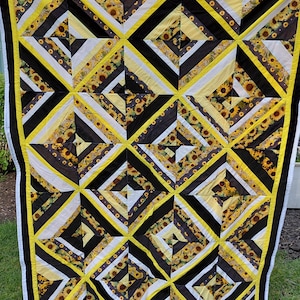 May include: A rectangular quilt featuring a geometric pattern of alternating diagonal stripes in black, white, and yellow. The fabric includes a sunflower print, solid black bands, and solid white bands, all framed by a bright yellow and black border.