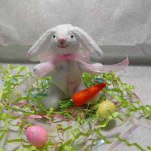 May include: A white plush bunny with a pink bow, sitting amidst Easter decorations. A bright orange carrot and two decorative eggs, one pink and one yellow, are also present. Green and pink paper shreds complete the festive scene.