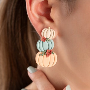 Può includere: Orecchini con un design a pila di zucche nei toni pastello pesca, azzurro e crema. Ogni zucca è decorata con piccoli dettagli floreali rossi, creando un accessorio stagionale.