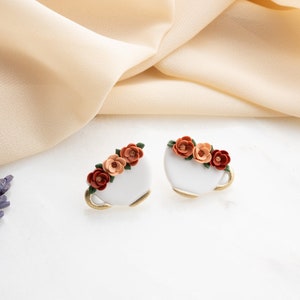 May include: A pair of white ceramic earrings shaped like teacups with a floral design. The flowers are orange, red, and green.