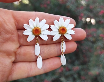 May include: A pair of white daisy earrings with gold chains and white petals dangling below. The daisies have orange centers.