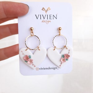 Pu&ograve; includere: Orecchini a forma di cuore bianchi con dettagli floreali rosa e verdi e cerchi dorati. Gli orecchini sono su una carta bianca con il testo "VIVIEN design" e "@viviendesign_"