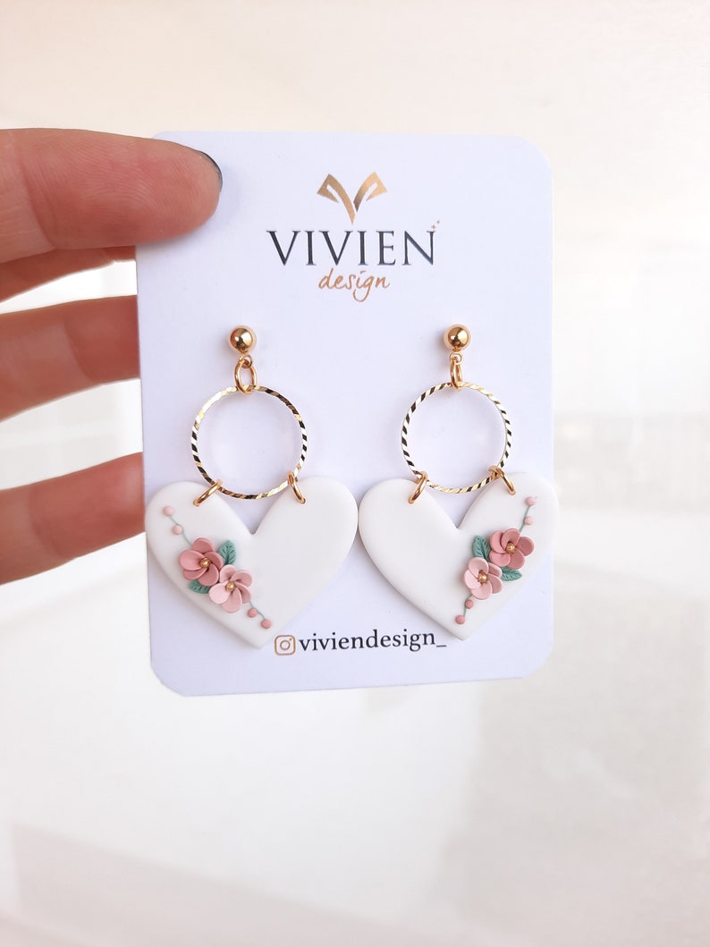 Pu&ograve; includere: Orecchini a forma di cuore bianchi con dettagli floreali rosa e verdi e cerchi dorati. Gli orecchini sono su una carta bianca con il testo "VIVIEN design" e "@viviendesign_"