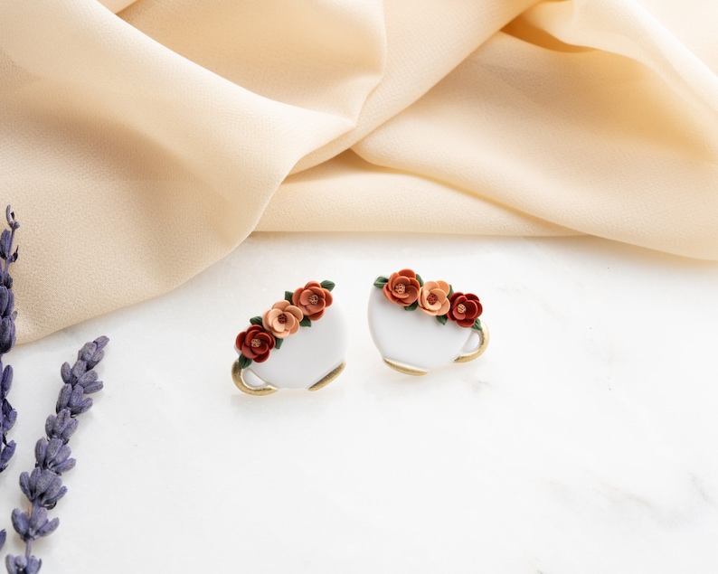 May include: A pair of white ceramic earrings shaped like teacups with a floral design. The flowers are orange, red, and green.