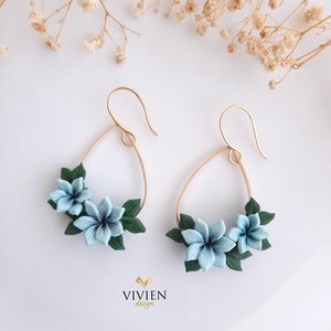 May include: A pair of gold hoop earrings with blue and green polymer clay flowers. The earrings are in the shape of a teardrop and have a delicate floral design.