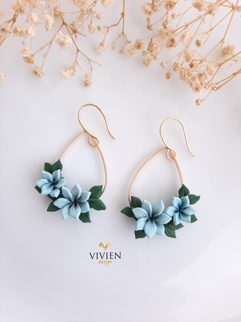 May include: A pair of gold hoop earrings with blue and green polymer clay flowers. The earrings are in the shape of a teardrop and have a delicate floral design.