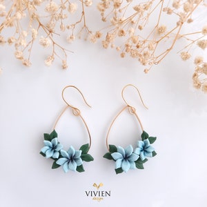 May include: A pair of gold hoop earrings with a teardrop shape. The hoops are adorned with three blue polymer clay flowers and green leaves.