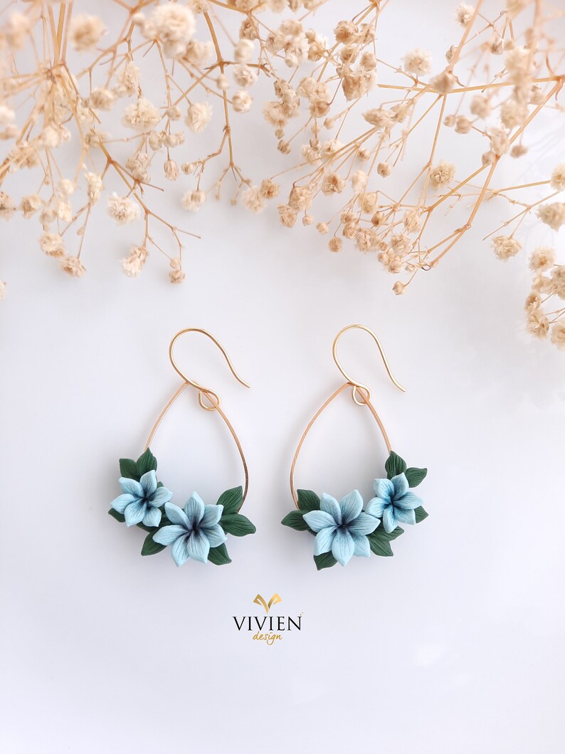 May include: A pair of gold hoop earrings with a teardrop shape. The hoops are adorned with three blue polymer clay flowers and green leaves.