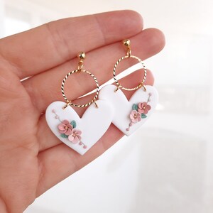 Pu&ograve; includere: Orecchini a forma di cuore bianchi con dettagli floreali rosa e verdi, appesi ad anelli dorati.