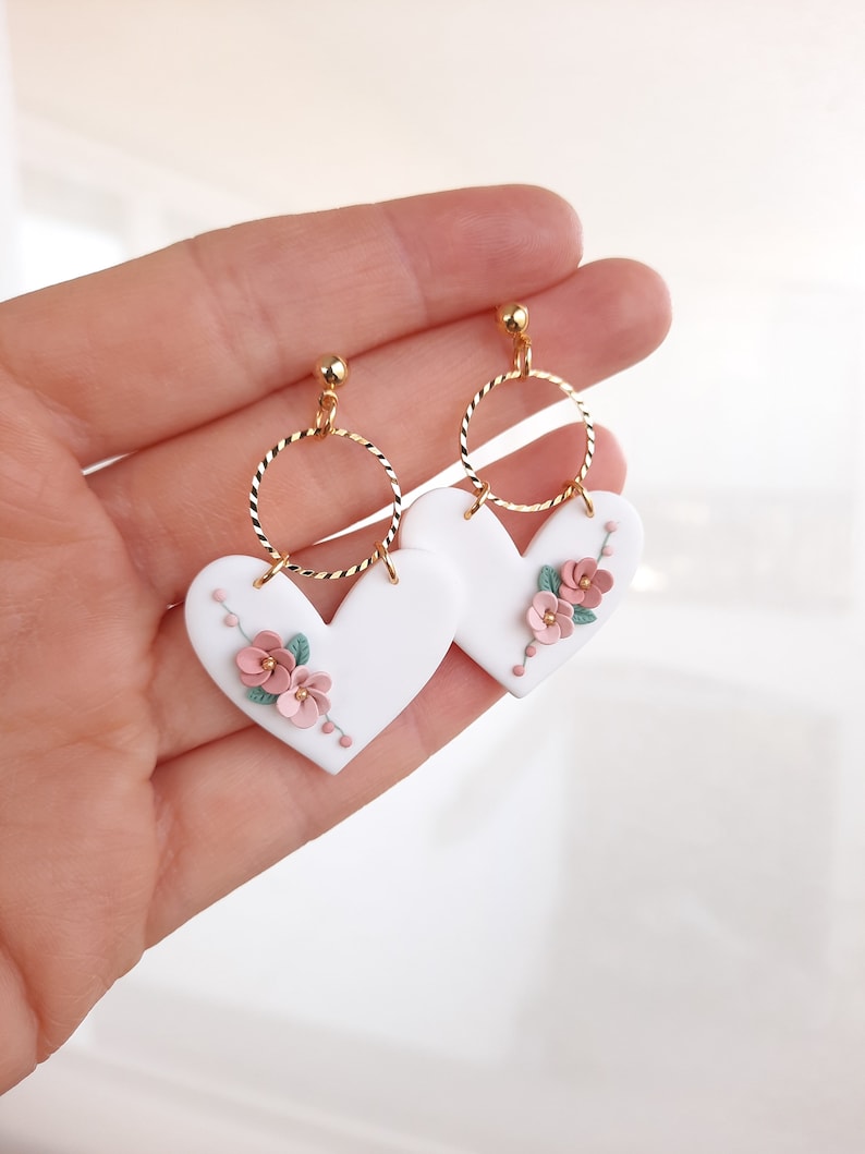 Pu&ograve; includere: Orecchini a forma di cuore bianchi con dettagli floreali rosa e verdi, appesi ad anelli dorati.