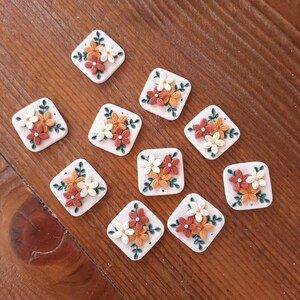 May include: Ten square-shaped ceramic tiles with a floral design. Each tile features a cluster of three flowers in shades of orange, white, and brown, with green leaves. The tiles have a white background and a slightly textured surface.