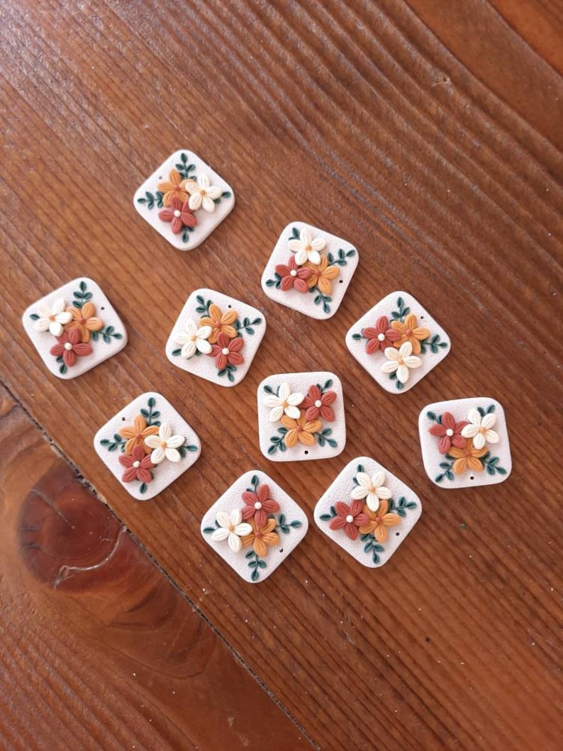 May include: Ten square-shaped ceramic tiles with a floral design. Each tile features a cluster of three flowers in shades of orange, white, and brown, with green leaves. The tiles have a white background and a slightly textured surface.