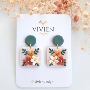 May include: A pair of floral earrings with a square base and a green circle charm. The earrings feature a white base with orange, yellow, and white flowers and green leaves.