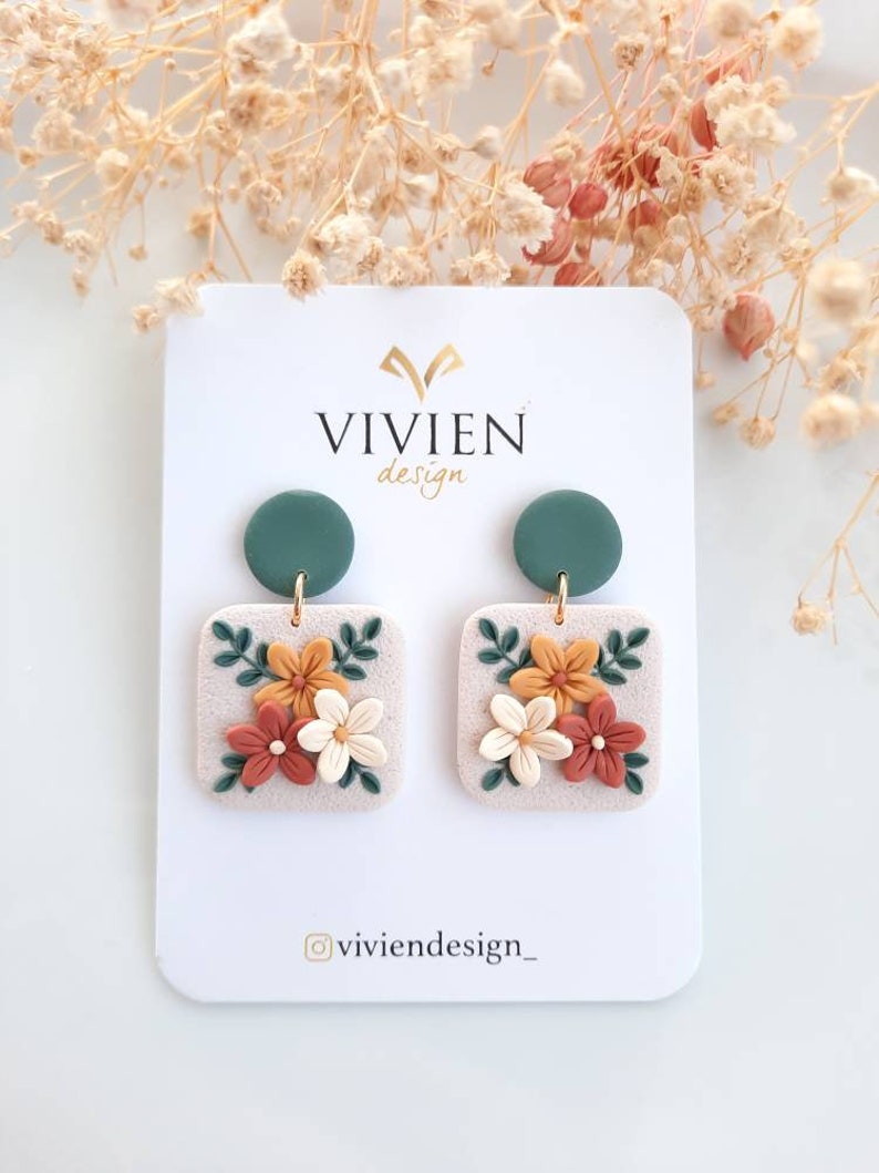 May include: A pair of floral earrings with a square base and a green circle charm. The earrings feature a white base with orange, yellow, and white flowers and green leaves.