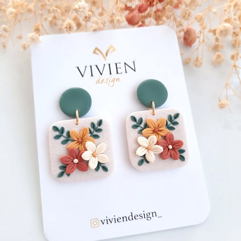 May include: A pair of dangle earrings with a square-shaped floral design. The earrings feature a light beige background with orange, white, and red flowers, green leaves, and a dark green circle at the top. The earrings are on a white card with the text "VIVIEN design" and "@viviendesign_" printed on it.