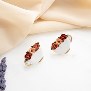 May include: A pair of white ceramic earrings shaped like teacups with a gold rim. Each earring is decorated with a cluster of orange, red, and peach colored flowers.