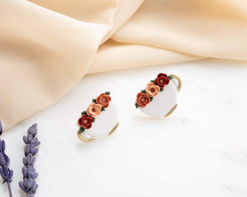 May include: A pair of white ceramic earrings shaped like teacups with a gold rim. Each earring is decorated with a cluster of orange, red, and peach colored flowers.