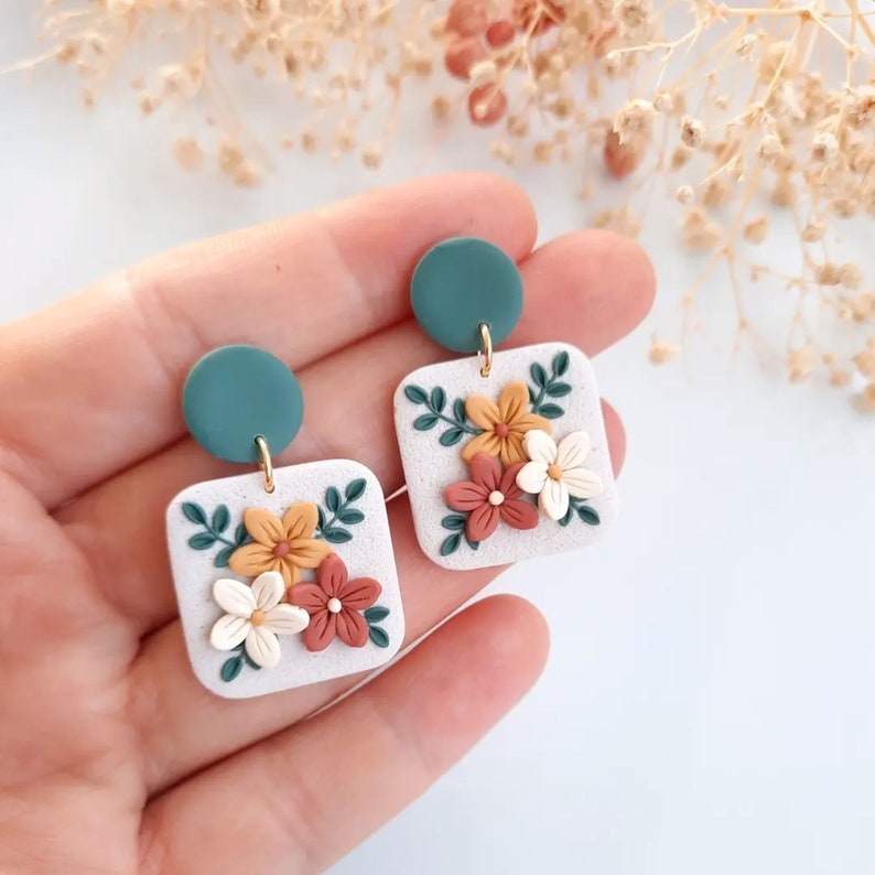 May include: A pair of square dangle earrings with a floral design. The earrings feature a white background with orange, white, and red flowers and green leaves. The top of each earring is a teal green circle.