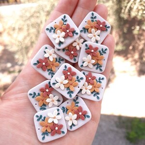 May include: A collection of 10 square-shaped polymer clay charms with floral designs. Each charm features a cluster of white, orange, and red flowers with green leaves on a white background.