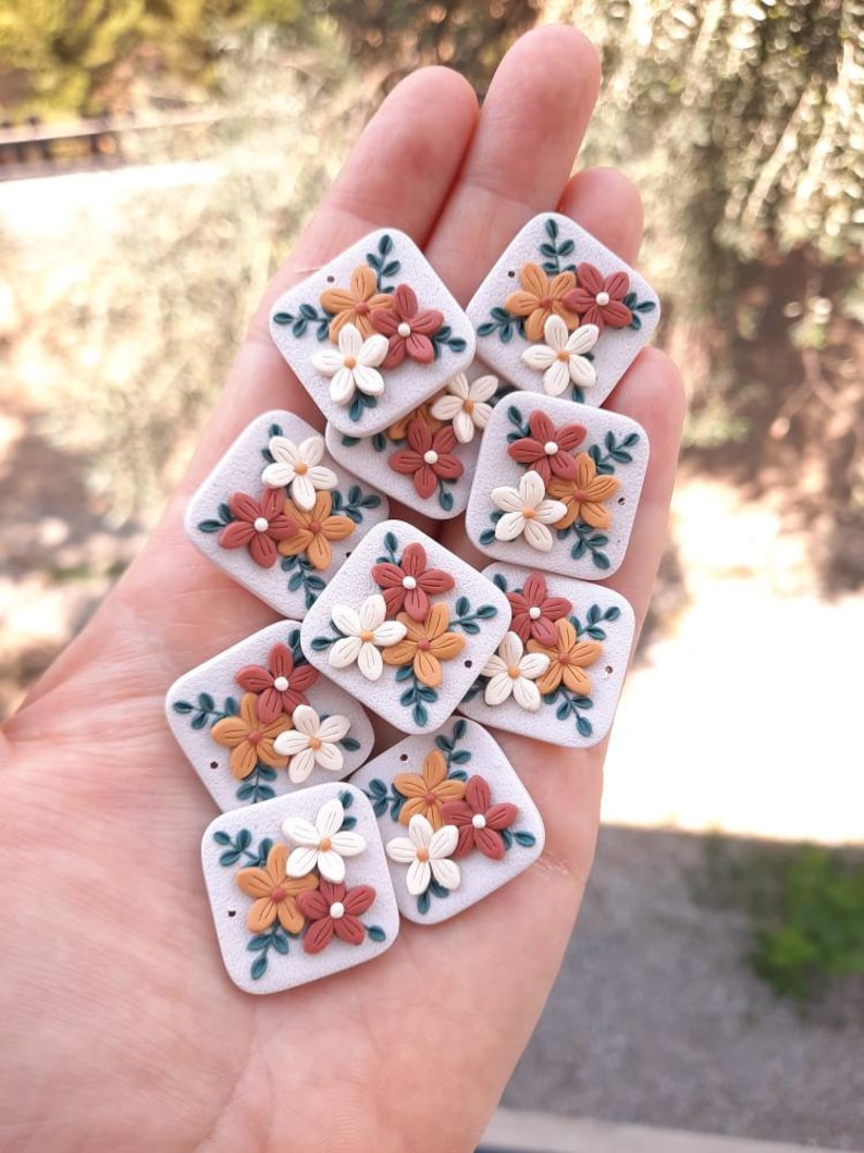 May include: A collection of 10 square-shaped polymer clay charms with floral designs. Each charm features a cluster of white, orange, and red flowers with green leaves on a white background.