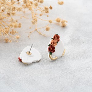 May include: A pair of white ceramic earrings with a floral design. The flowers are red, orange, and green. The earrings are stud style with a post and a butterfly backing.