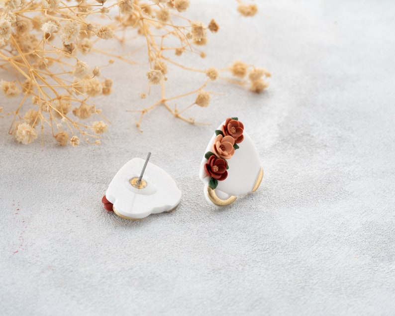 May include: A pair of white ceramic earrings with a floral design. The flowers are red, orange, and green. The earrings are stud style with a post and a butterfly backing.