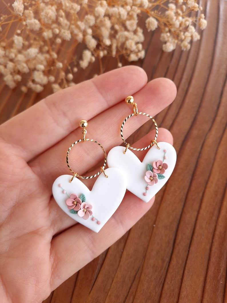 Pu&ograve; includere: Orecchini a forma di cuore bianchi con delicati accenti floreali rosa e verdi. Gli orecchini hanno un cerchio dorato e un perno dorato.