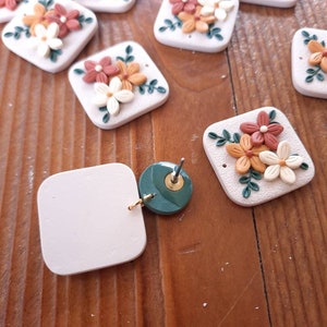 May include: A set of handmade polymer clay earrings featuring floral designs with white, orange, and brown accents. The earrings are in the shape of a square and have a green leaf design. The earrings are shown on a wooden surface.