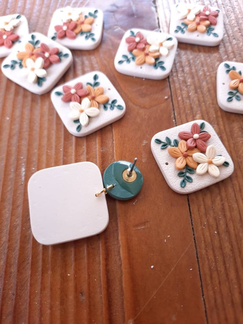 May include: A set of handmade polymer clay earrings featuring floral designs with white, orange, and brown accents. The earrings are in the shape of a square and have a green leaf design. The earrings are shown on a wooden surface.