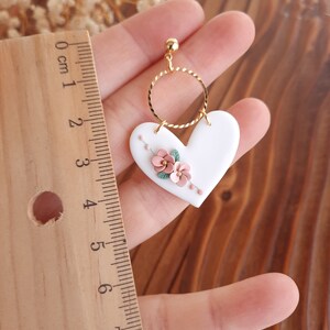Pu&ograve; includere: Orecchini in ceramica bianca a forma di cuore con un cerchio di filo d'oro e accenti floreali rosa.