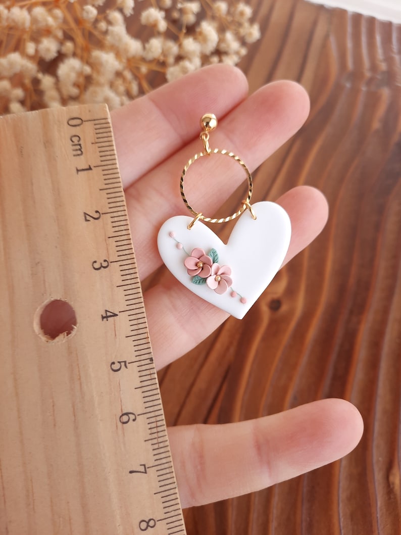 Pu&ograve; includere: Orecchini in ceramica bianca a forma di cuore con un cerchio di filo d'oro e accenti floreali rosa.