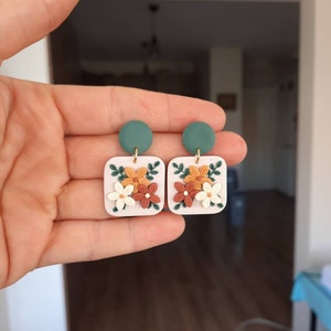 May include: A pair of square-shaped earrings with a floral design. The earrings are made of white clay with orange, brown, and green flowers. The earrings have a green circle at the top.