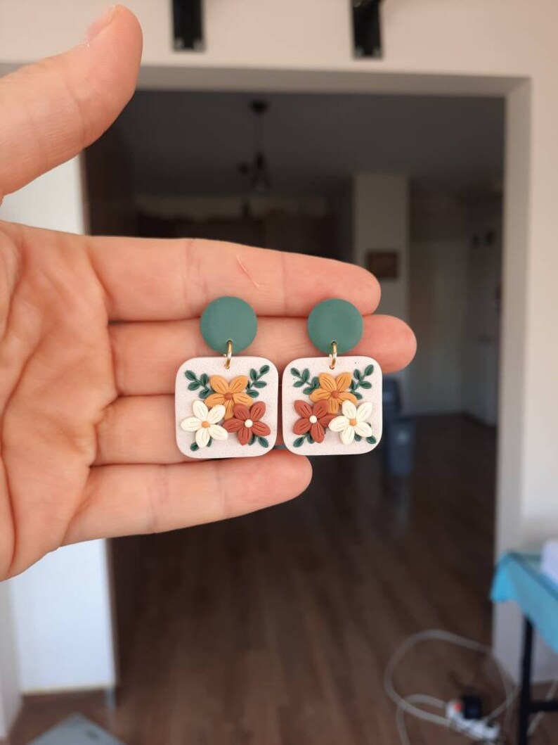 May include: A pair of square-shaped earrings with a floral design. The earrings are made of white clay with orange, brown, and green flowers. The earrings have a green circle at the top.