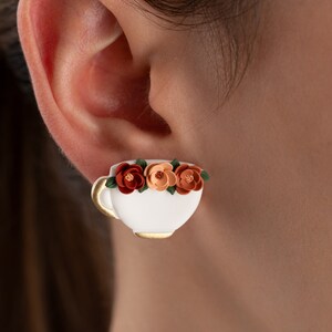 May include: White ceramic teacup earrings with a gold rim and three orange and brown flowers.