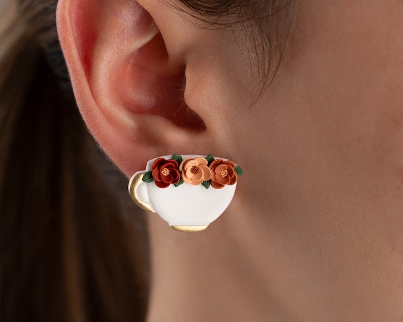 May include: White ceramic teacup earrings with a gold rim and three orange and brown flowers.