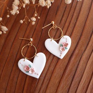 Pu&ograve; includere: Un paio di orecchini a forma di cuore bianco con delicati fiori rosa e foglie verdi. Gli orecchini hanno un cerchio dorato e un perno dorato.