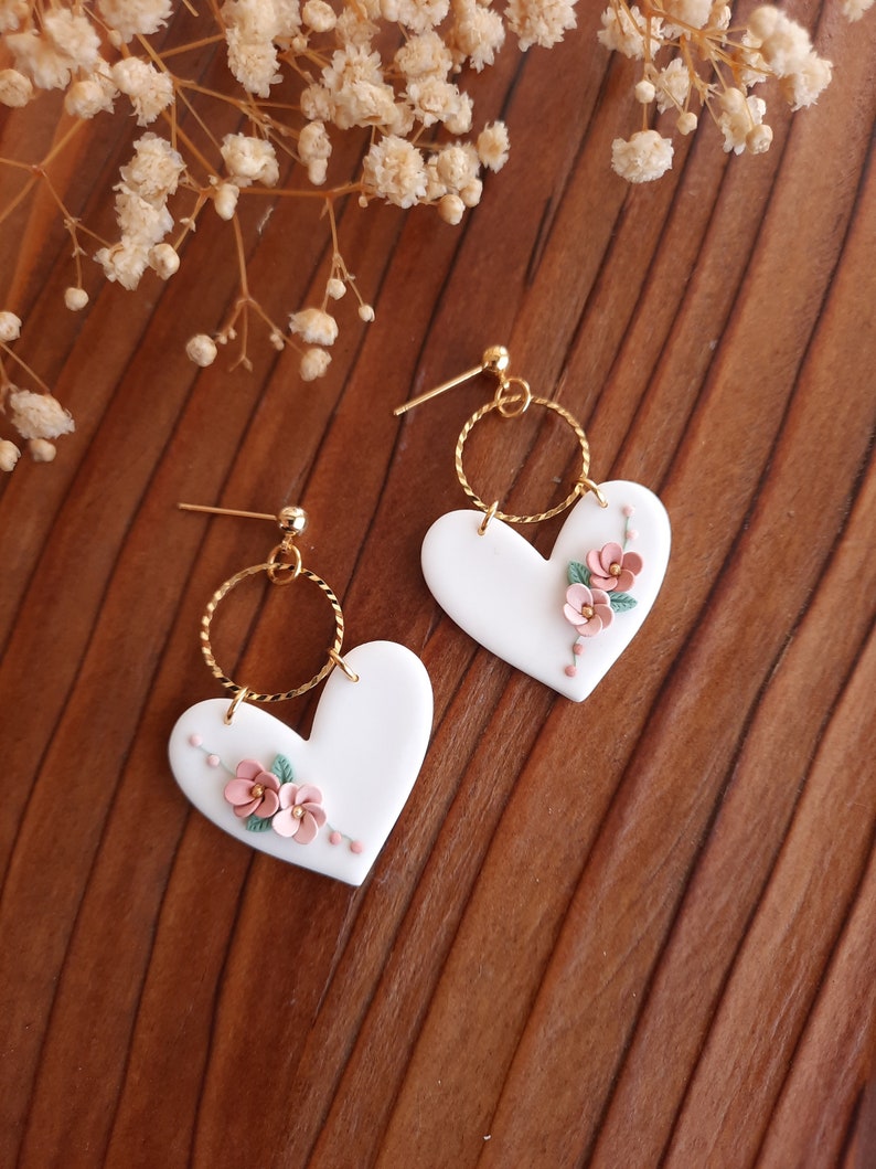 Pu&ograve; includere: Un paio di orecchini a forma di cuore bianco con delicati fiori rosa e foglie verdi. Gli orecchini hanno un cerchio dorato e un perno dorato.