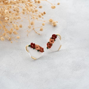 May include: A pair of white ceramic earrings with a gold rim and a floral design. The flowers are orange, red, and green.