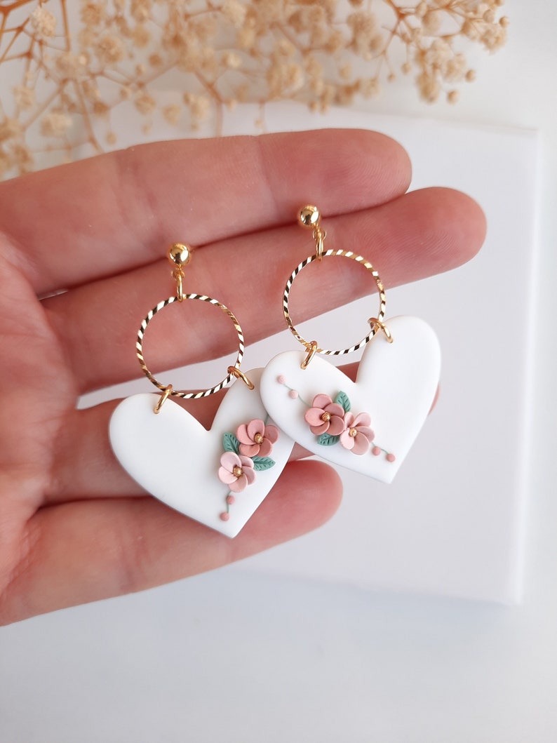 Pu&ograve; includere: Orecchini a forma di cuore bianchi con cerchi dorati e dettagli floreali rosa e verdi.