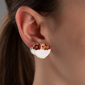May include: White ceramic teacup earring with a gold rim and a floral design of orange, peach, and green flowers.