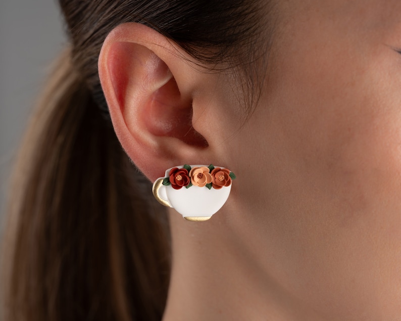 May include: White ceramic teacup earring with a gold rim and a floral design of orange, peach, and green flowers.