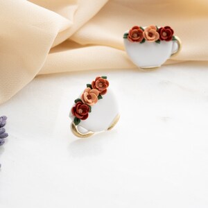 May include: A pair of white ceramic teacups with gold trim, each decorated with a cluster of orange, red, and brown flowers.