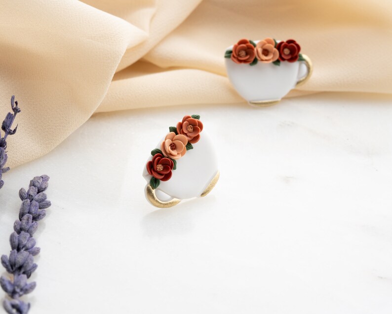 May include: A pair of white ceramic teacups with gold trim, each decorated with a cluster of orange, red, and brown flowers.