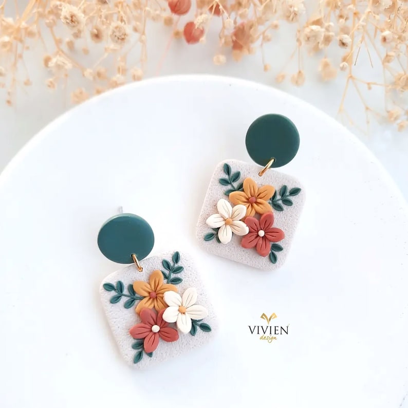 May include: A pair of floral earrings with a square-shaped base and a green circle charm. The earrings feature a white, orange, and red floral design with green leaves.