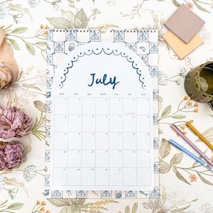 May include: A July calendar with a blue and white decorative border, displayed on a floral patterned surface. The calendar is accompanied by pens, sticky notes, a mug, and flowers. The word "July" is written in blue script.
