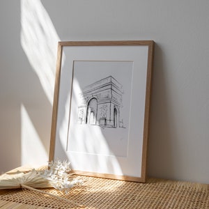 May include: A framed black and white line drawing of the Arc de Triomphe in Paris, France. The drawing is on white paper and is framed in a natural wood frame.