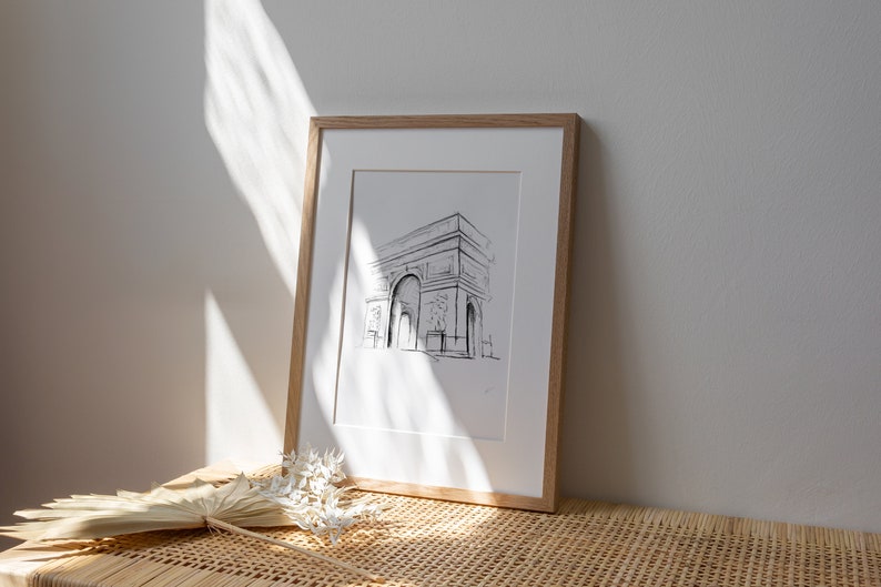 May include: A framed black and white line drawing of the Arc de Triomphe in Paris, France. The drawing is on white paper and is framed in a natural wood frame.