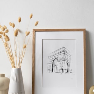 May include: A framed black and white line drawing of the Arc de Triomphe in Paris, France. The drawing is in a simple, minimalist style. The frame is made of light brown wood.