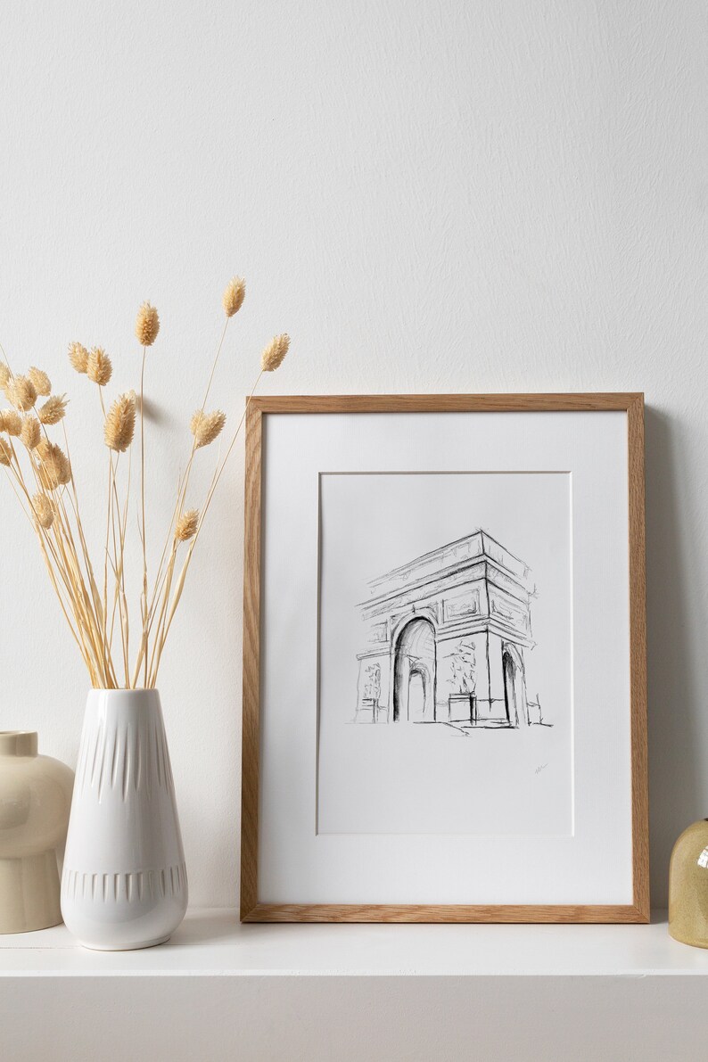 May include: A framed black and white line drawing of the Arc de Triomphe in Paris, France. The drawing is in a simple, minimalist style. The frame is made of light brown wood.