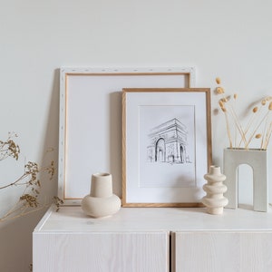 May include: A white cabinet with two doors, a framed black and white drawing of the Arc de Triomphe, a white vase, a beige vase, and dried flowers in a gray vase.